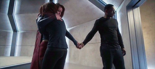 A scene from Supergirl: Kara holds her sobbing sister, Alex, with one arm while holding hands with their father-figure, J'onn, with the other as he looks away. 